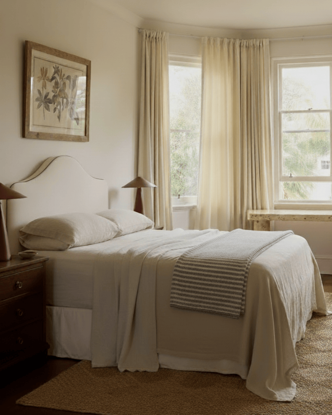Neatly made bed in a bedroom with curtains and a painting on the wall.