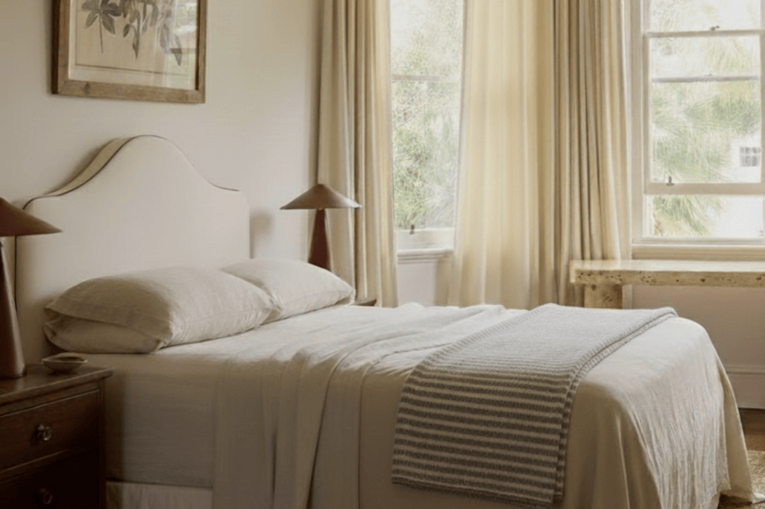 Neatly made bed in a bedroom with curtains and a painting on the wall.