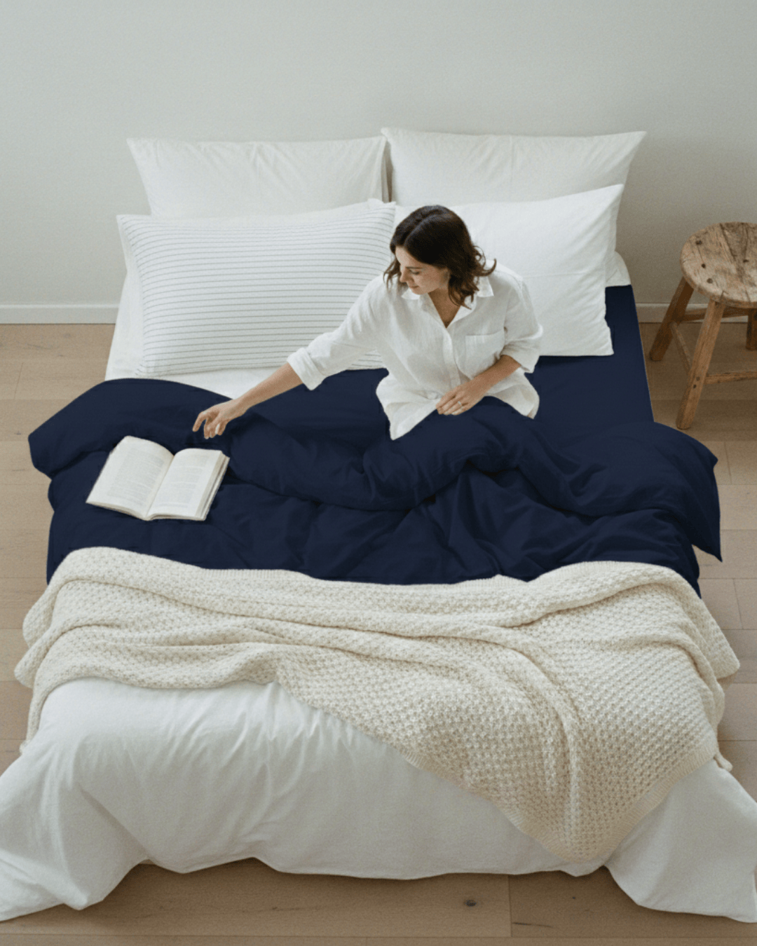 Woman sitting on a bed with navy bedding and white pillows, reading a book.