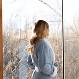 Woman in a blue robe standing by a window with a snowy landscape outside.