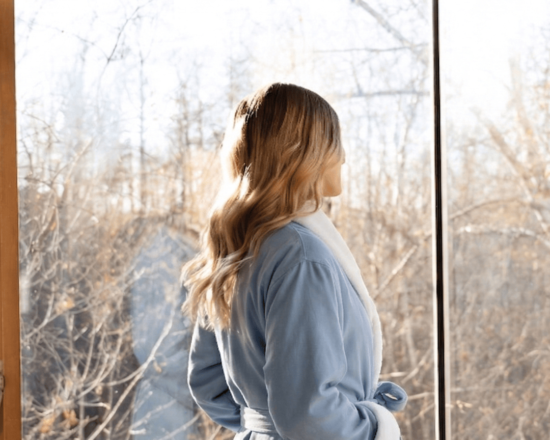 Woman in a blue robe standing by a window with a snowy landscape outside.