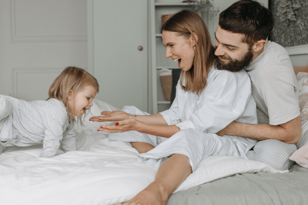Family of three in a bedroom setting, with a child lying on the bed and parents sitting close by.