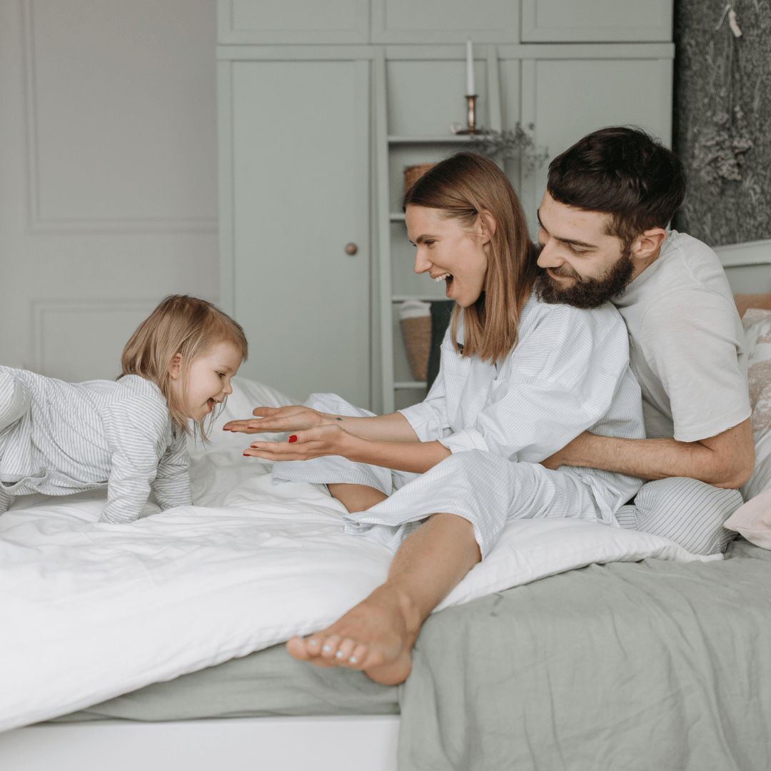 Family of three in a bedroom setting, with a child lying on the bed and parents sitting close by.