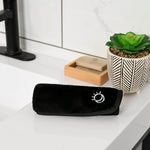 Glow Away Makeup remover cloth in Black towel with Skylark+Owl logo on a white surface with a plant and soap in the background