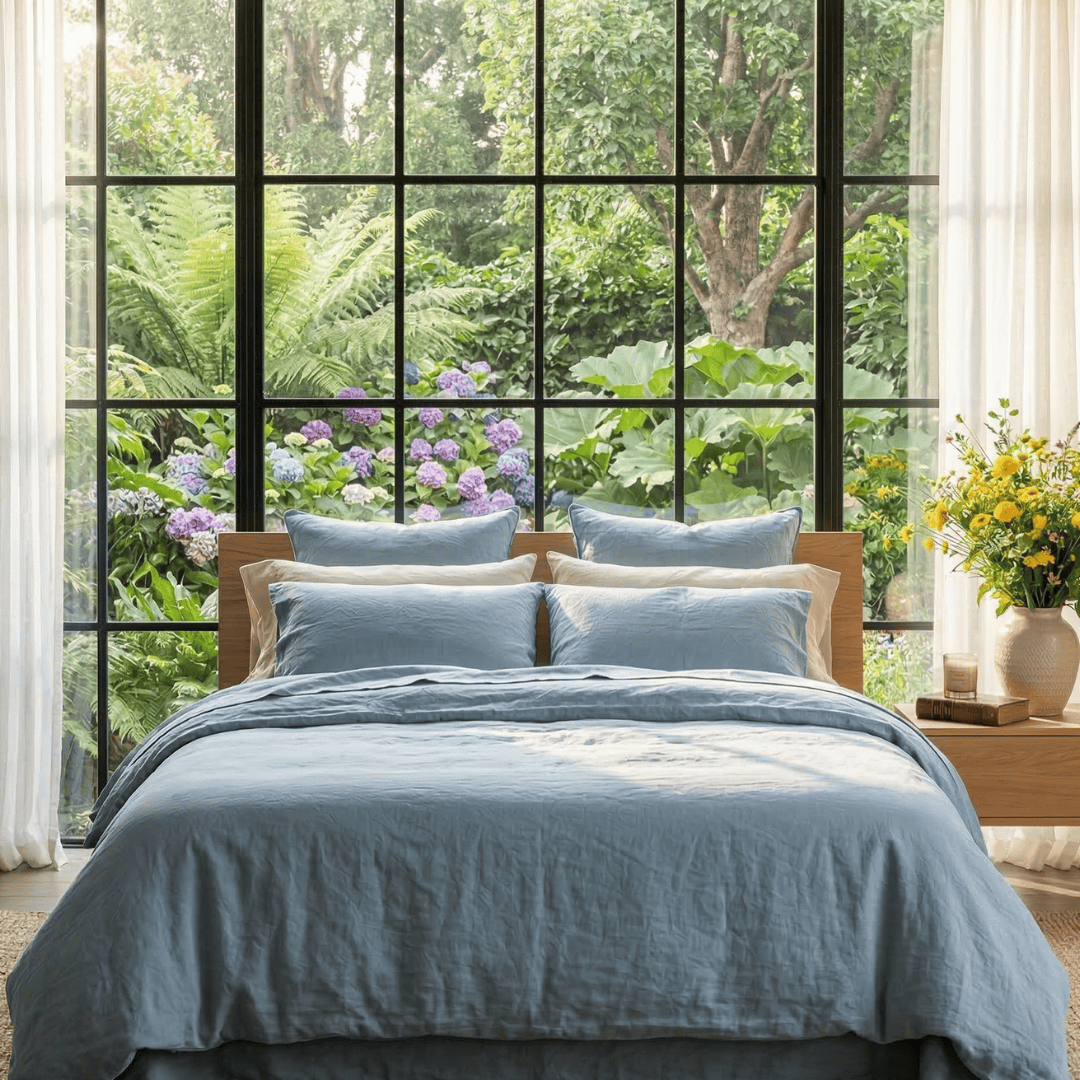 Bedroom with a large window view of greenery and flowers, featuring a bed with blue bedding.
