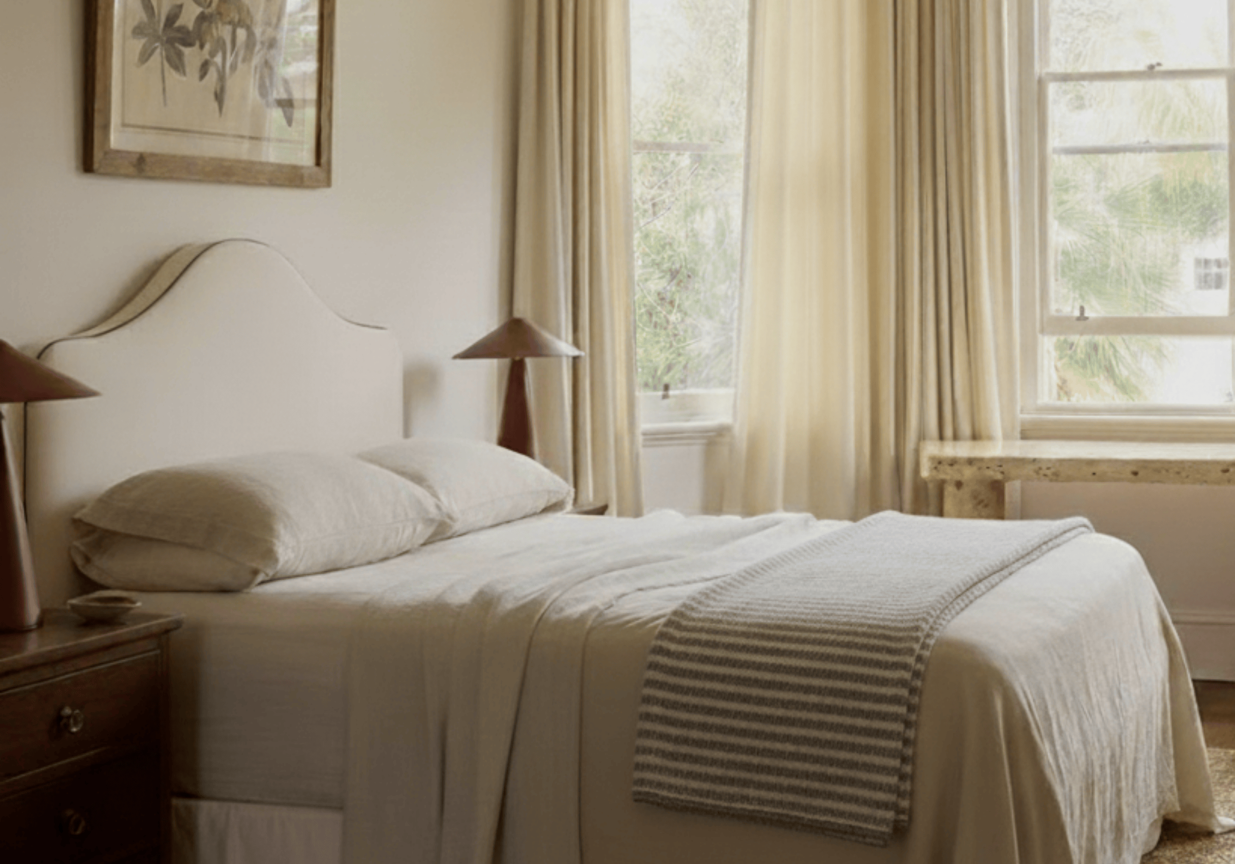 Neatly made bed in a bedroom with curtains and a painting on the wall.