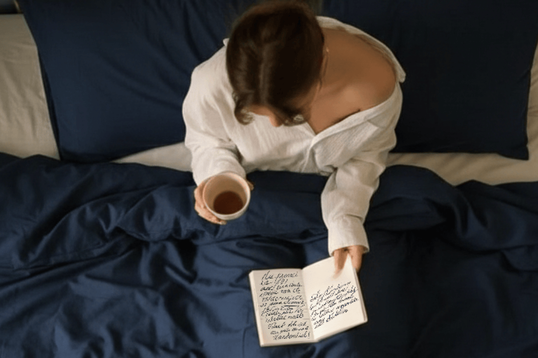 Person sitting on a bed holding a cup and a notebook with a dark blue comforter.