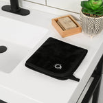 Black towel with a logo on a kitchen counter next to a sink, soap, and plant.
