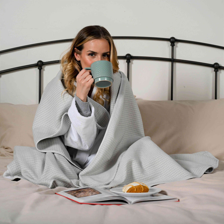 Woman wrapped in Earl Grey Brushed Cotton Blanket, sitting on a bed while sipping a warm drink